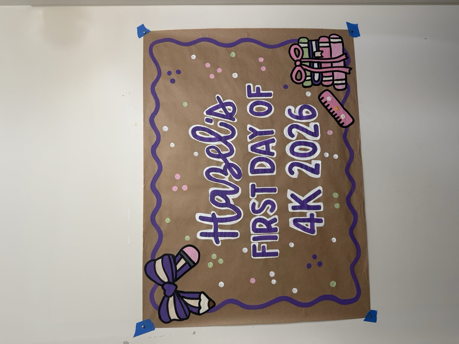 Brown paper banner reading Hazel's First Day of 4K 2026 in purple bubble letters, framed by a purple wavy border with a painted bow, ruler and stack of books.