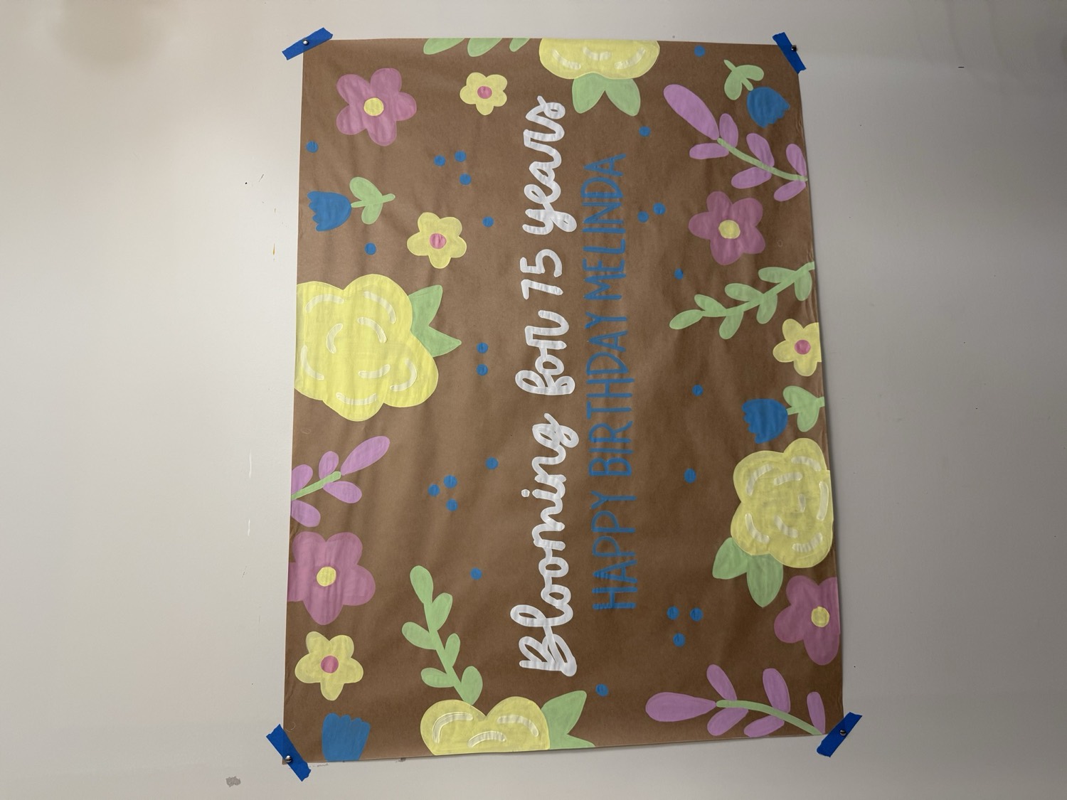 Brown paper banner reading Blooming for 75 Years, Happy Birthday Melinda, surrounded by hand-painted pink, yellow and blue flowers with green leaves.