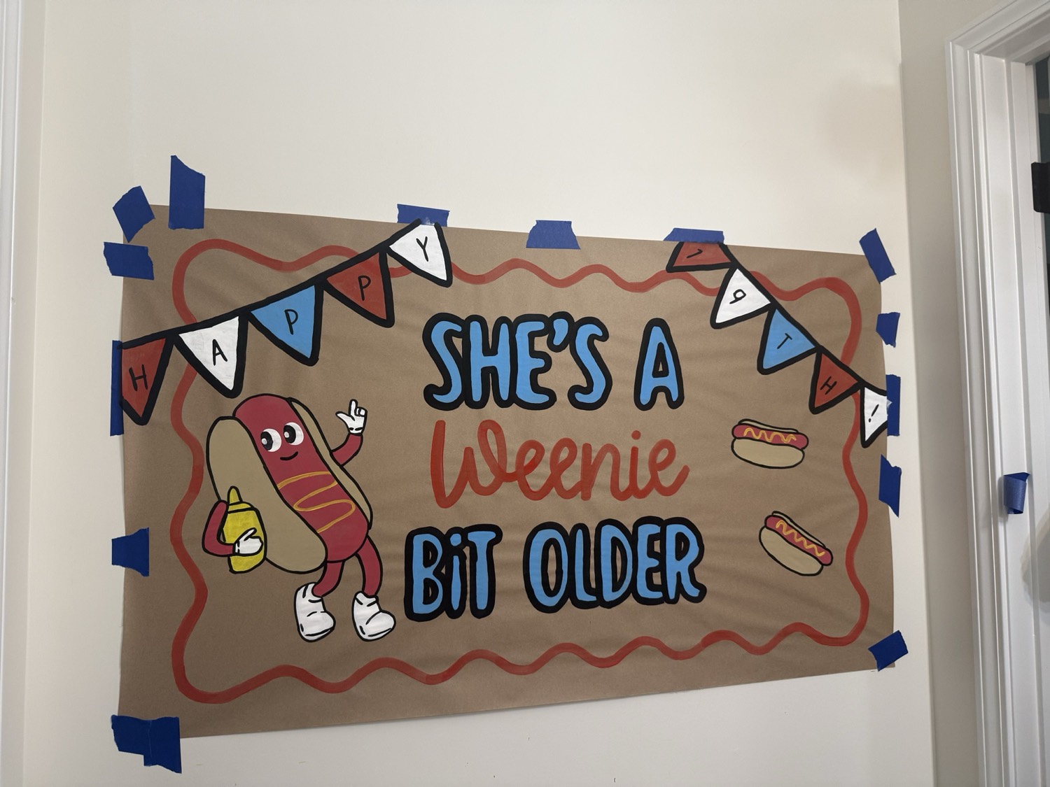 Brown paper banner reading She's a Weenie Bit Older, with a smiling cartoon hot dog holding a mustard bottle and a red, white and blue pennant garland across the top.