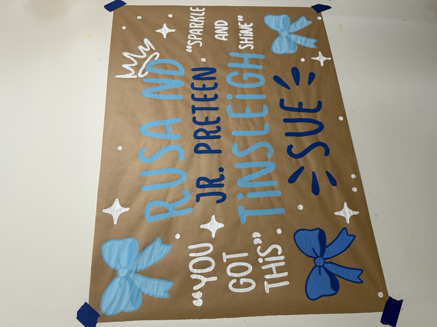 Brown paper banner reading Rusa ND Jr Preteen Tinsleigh Sue in two shades of blue, with painted bows, sparkles, a crown and the phrases You Got This and Sparkle and Shine.