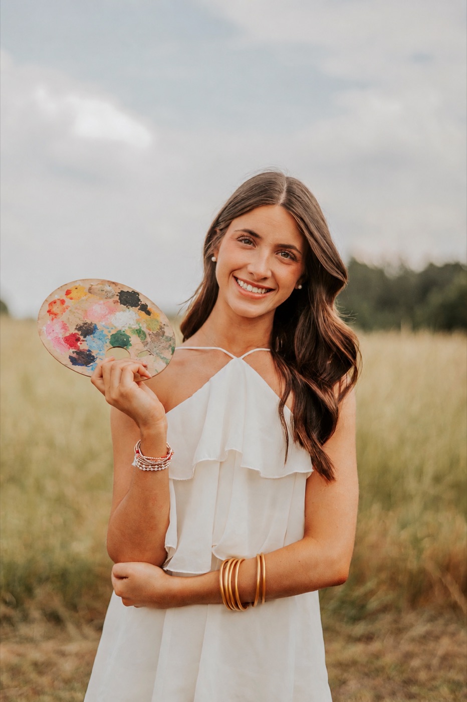 Kennedy standing in a field in a white dress, smiling and holding a well-used paint palette.