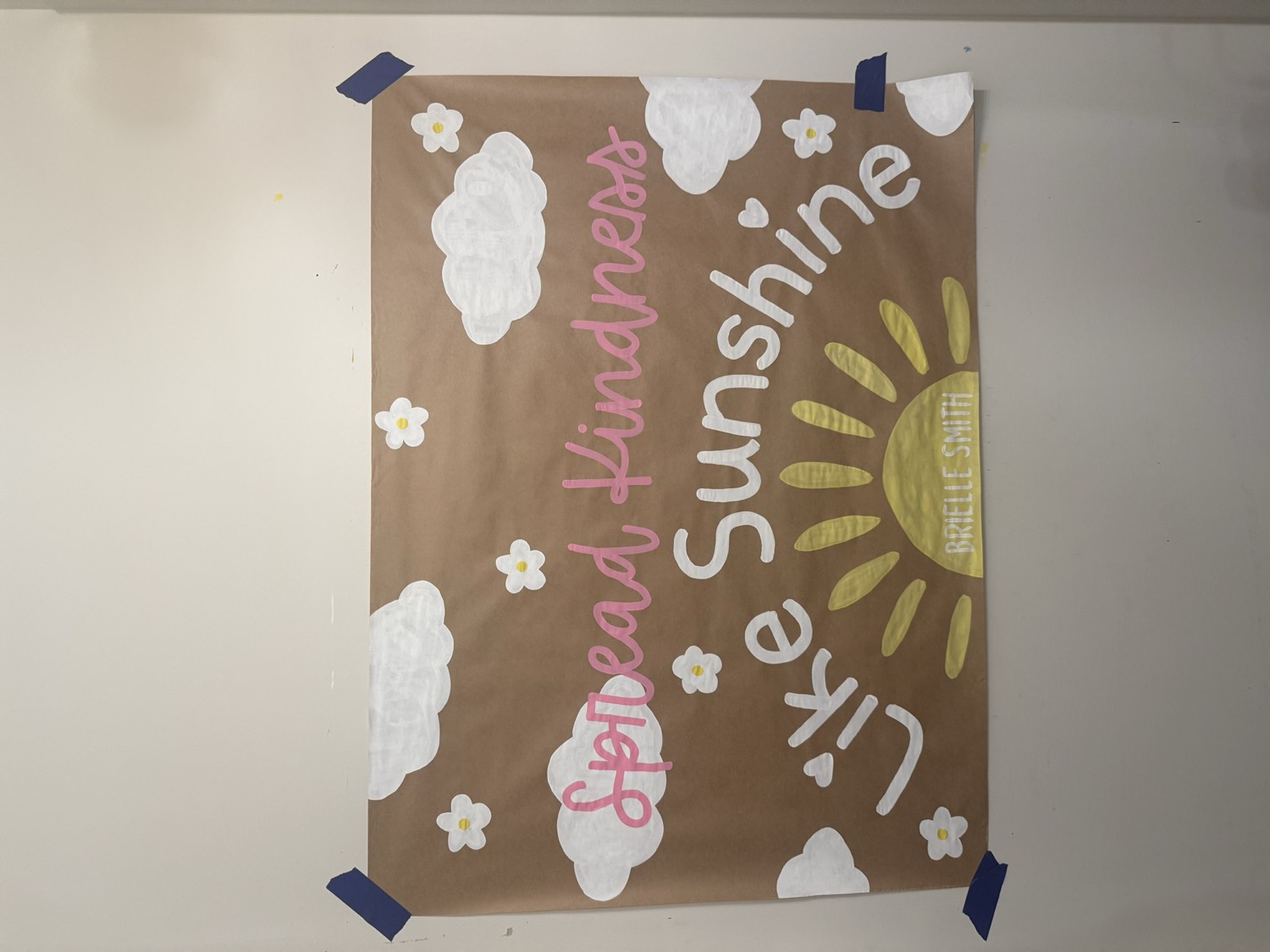 Brown paper banner with Spread Kindness Like Sunshine in pink and white lettering above a big yellow sun, surrounded by painted clouds and daisies.