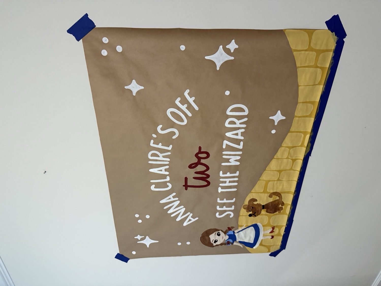 Brown paper banner reading Anna Claire's Off Two See The Wizard, with Dorothy and Toto painted at the end of a yellow brick road.